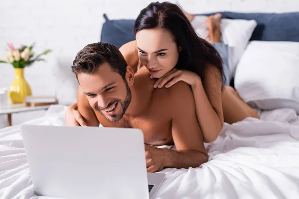 Sexy brunette woman lying on shirtless man smiling and looking at laptop on bed on blurred background — Stock Photo