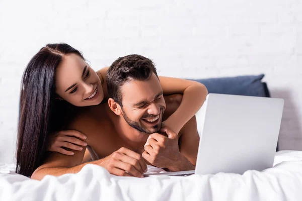 Счастливая брюнетка обнимает мужчину без рубашки, смеющегося возле ноутбука на кровати — стоковое фото