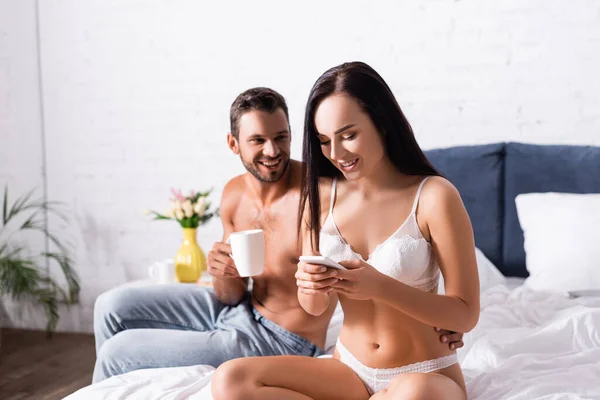 Sorrindo homem com xícara de café abraçando mulher sexy em lingerie, mensagens de texto no smartphone no fundo borrado no quarto — Fotografia de Stock