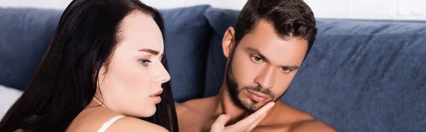 Surprised brunette woman looking away while touching face of displeased man in bedroom, banner — Stock Photo