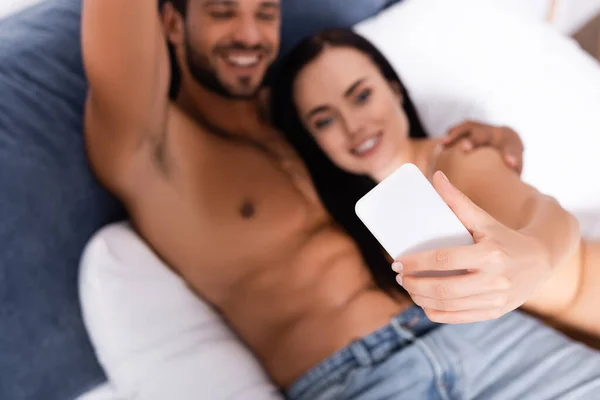 Feliz jovem casal com smartphone tomando selfie enquanto deitado na cama no fundo borrado — Fotografia de Stock