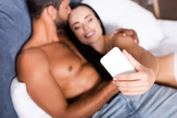 Shirtless man kissing happy woman on bed during selfie on blurred background — Stock Photo