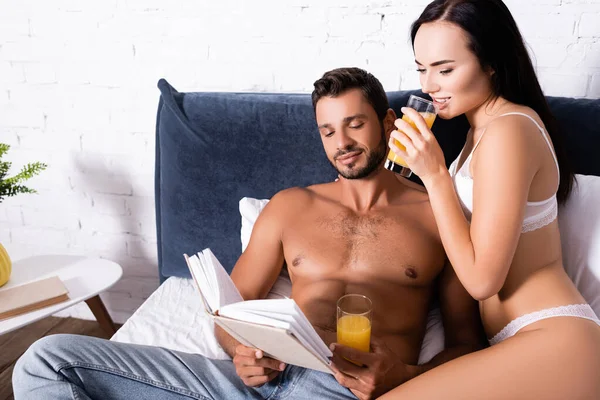 Sonriente hombre sin camisa leyendo libro cerca de mujer en lencería bebiendo jugo en la cama - foto de stock