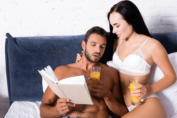 Sexy joven pareja con vasos de jugo lectura libro en la cama - foto de stock