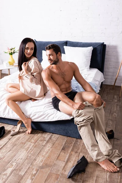 Toute la longueur de sourire jeune couple portant tout en étant assis sur le lit — Photo de stock