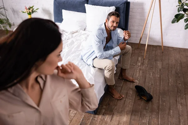 Smiling young man looking at woman while wearing on bed on blurred foreground in bedroom — Stock Photo