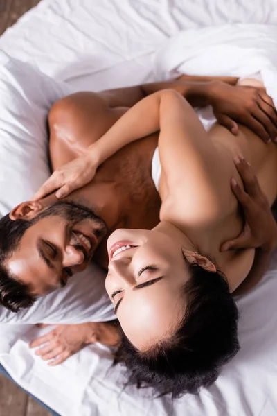 Vista ad alto angolo di felice giovane uomo abbracciando donna sensuale con gli occhi chiusi sul letto — Foto stock