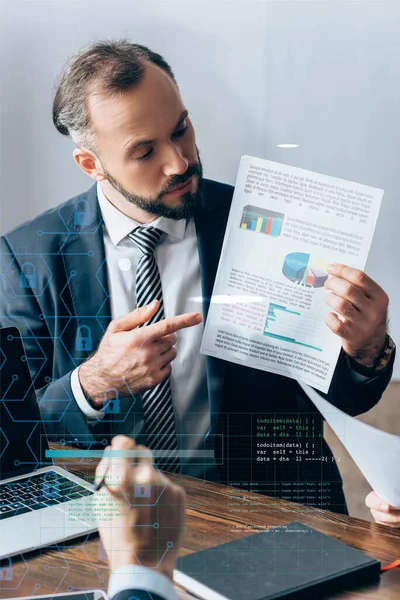 Businessman pointing at paper with charts near laptop, colleague and investor on blurred foreground, illustration of cyber font — Stock Photo