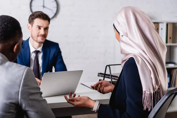 Back view of arabian businesswoman gesturing while talking to european and african american partners, blurred background — Stock Photo