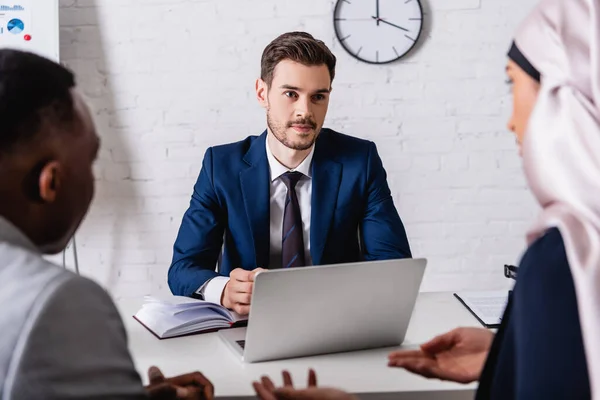 European translator sitting near laptop and looking at arabian and african american business partners, blurred foreground — Stock Photo