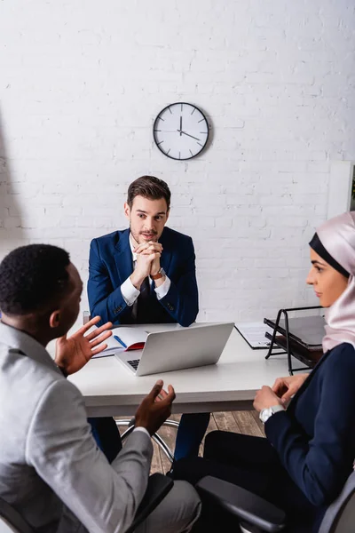 Homme d'affaires afro-américain gestuelle tout en parlant à des partenaires d'affaires multiculturels lors de la réunion, flou premier plan — Photo de stock