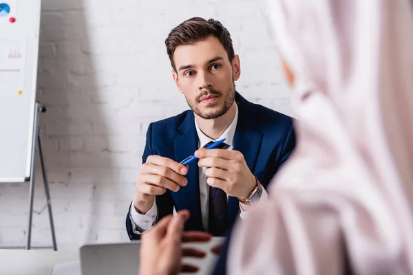 Seriöser Übersetzer hält Stift in der Hand, während er arabische Geschäftsfrau im verschwommenen Vordergrund ansieht — Stockfoto