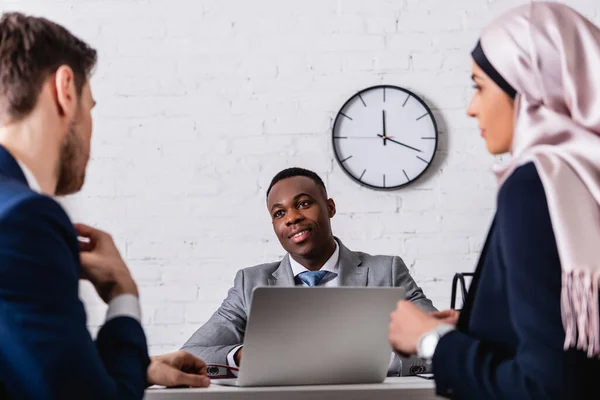 Sonriente hombre de negocios afroamericano mirando al traductor cerca de la mujer de negocios árabe en el cargo, borrosa primer plano - foto de stock