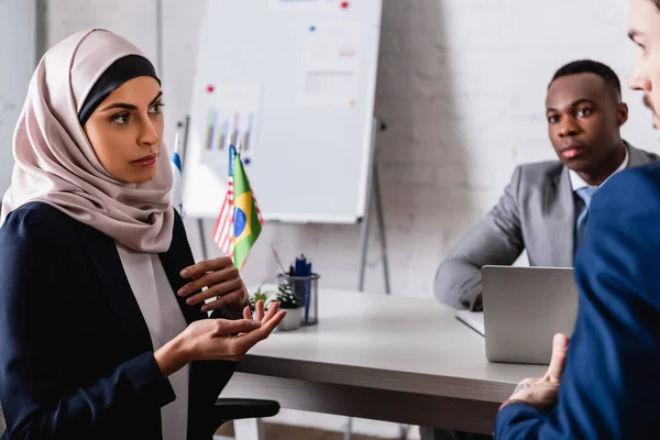 Femme d'affaires arabe dans le hijab geste tout en parlant à des partenaires multiculturels lors de la réunion, flou premier plan — Photo de stock