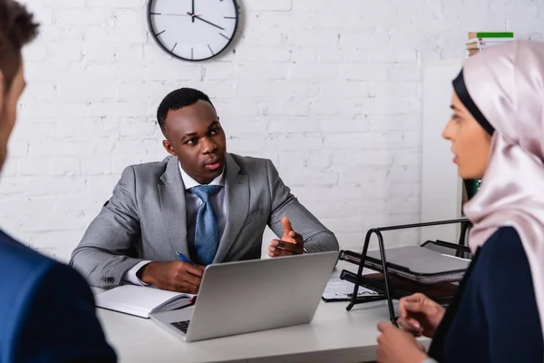 Afrikanisch-amerikanischer Geschäftsmann gestikuliert im Gespräch mit arabischer Geschäftsfrau in der Nähe eines Übersetzers, verschwommener Vordergrund — Stockfoto