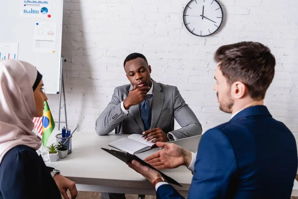 Dolmetscher zeigt mit der Hand auf Klemmbrett in der Nähe arabischer und afrikanisch-amerikanischer Geschäftspartner im unscharfen Vordergrund — Stockfoto