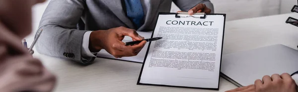 Vista recortada del hombre de negocios afroamericano apuntando con la pluma en el contrato cerca de socios comerciales en primer plano borroso, bandera - foto de stock