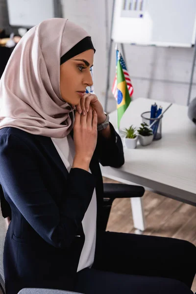 Thoughtful arabian businesswoman in hijab sitting in office near international flags on blurred background — Stock Photo