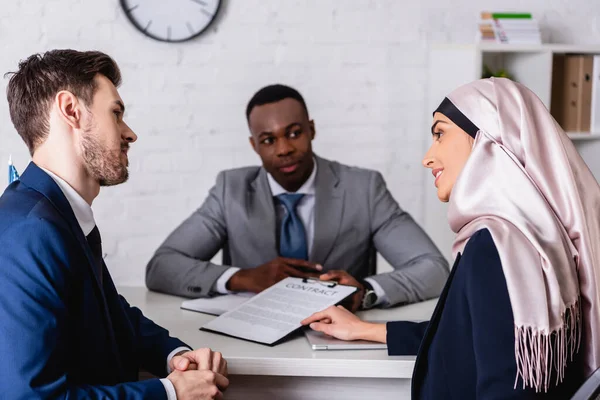 Sorridente empresária árabe apontando com a mão no contrato durante discussão com parceiros de negócios multiétnicos, fundo turvo — Fotografia de Stock
