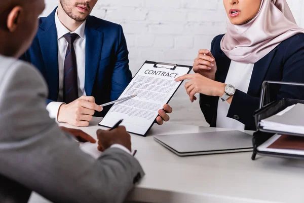 Partial view of translator pointing at contract near arabian and african american business partners, blurred foreground — Stock Photo