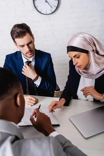 Mujer de negocios árabe señalando con el dedo en el contrato durante la reunión con el socio afroamericano y el intérprete, borrosa primer plano - foto de stock