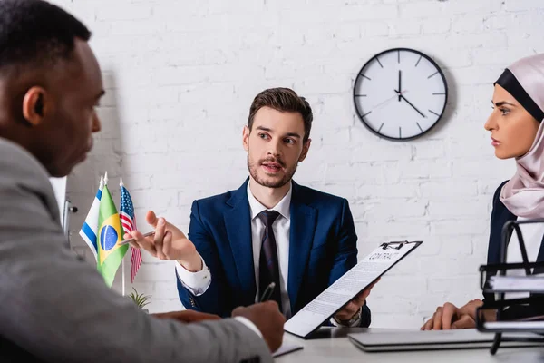 Businessman holding contract and gesturing during negotiation with interracial business partners, blurred foreground — Stock Photo