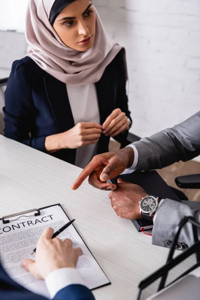 Arabian businesswoman looking at african american businessman pointing with finger at contract near translator, blurred foreground — Stock Photo