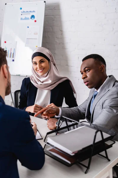 Homme d'affaires afro-américain pointant du doigt une femme d'affaires arabique souriante et traductrice au premier plan flou — Photo de stock