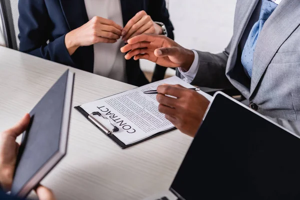 Cropped view of african american businessman holding pen and gesturing near contract and interracial business partners, blurred foreground — Stock Photo