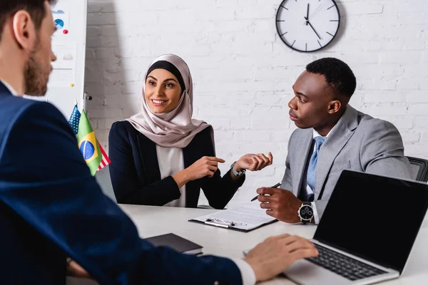 Smiling arabian businesswoman pointing with hands during meeting with multicultural business partners, blurred foreground — Stock Photo