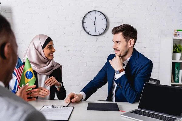 Alegre mujer de negocios árabe sonriendo durante el encuentro con socios de negocios multiculturales, borrosa primer plano - foto de stock