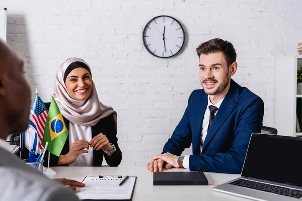 Alegres socios de negocios multiculturales sonriendo mientras miran al hombre de negocios afroamericano en primer plano borroso - foto de stock