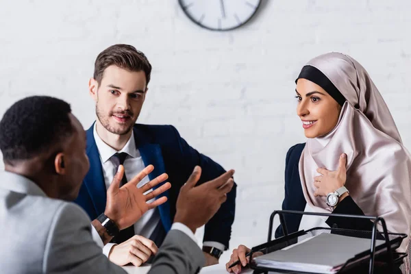 Empresario afroamericano gesticulando durante la discusión con sonriente socio de negocios multicultural e intérprete, borrosa primer plano - foto de stock