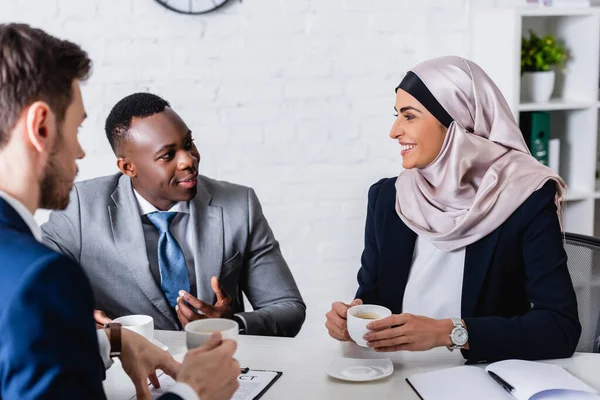 Sorridente empresário afro-americano gesticulando perto de empresário afro-americano e intérprete segurando xícaras de café, foreground borrado — Fotografia de Stock