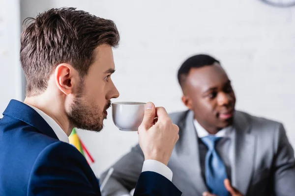 Intérprete beber café perto de parceiro de negócios afro-americano em fundo turvo — Fotografia de Stock