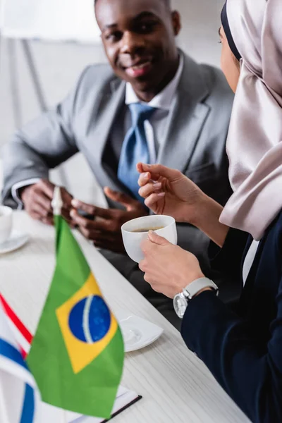 Selective focus of arabian businessman holding cup of coffee near brazilian flag and african american business partner on blurred background — Stock Photo