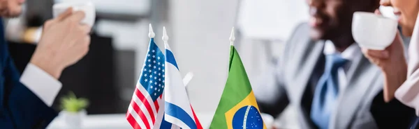 Selective focus of american, brazilian and usa flags near multicultural businesspeople drinking coffee on blurred background, partial view, banner — Stock Photo