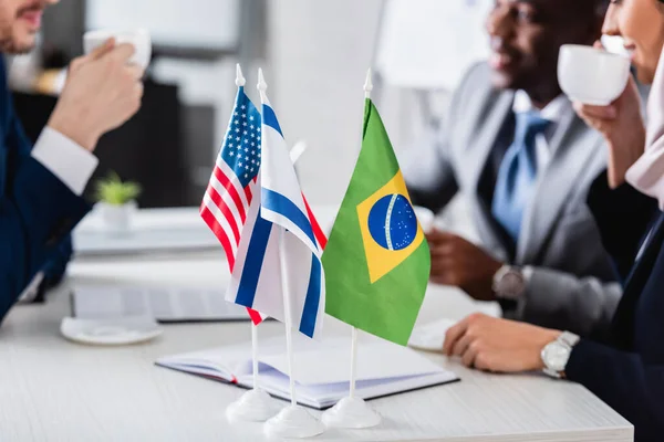 Selektiver Fokus amerikanischer, brasilianischer und israelischer Flaggen in der Nähe von interrassischen Geschäftspartnern, die Kaffee mit Dolmetscher trinken, beschnittene Ansicht — Stockfoto