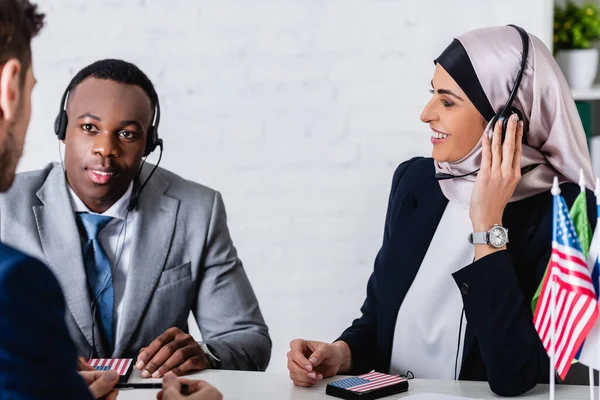 Sonrientes socios comerciales árabes y afroamericanos en auriculares cerca de intérprete en primer plano borroso - foto de stock