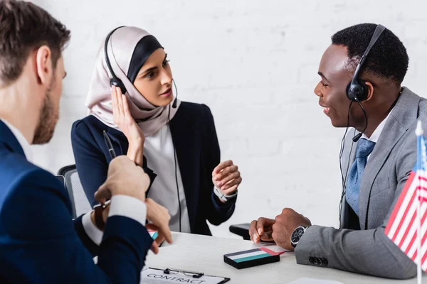 Arabian and african american business partners in headsets near translator on blurred foreground — Stock Photo