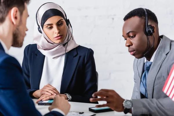 Arabian and african american business people in headsets near digital translators and interpreter on blurred foreground — Stock Photo