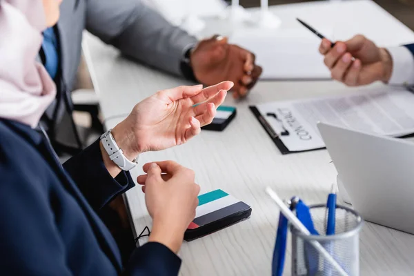 Vista recortada de la mujer de negocios árabe señalando con la mano cerca de socios comerciales multiculturales y traductores digitales, fondo borroso - foto de stock