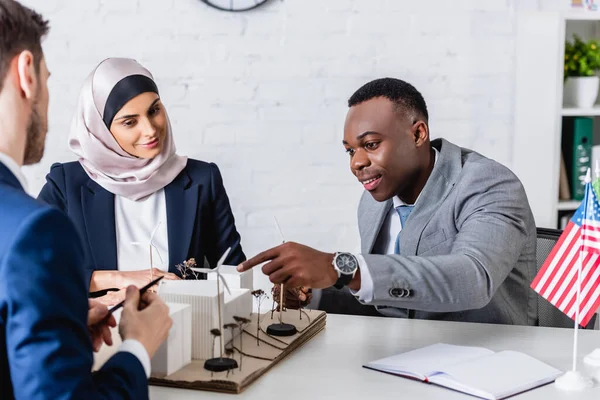 Felice uomo d'affari afroamericano che punta al modello di centrale elettrica alternativa con turbine eoliche vicino a partner multietnici, in primo piano sfocato — Foto stock