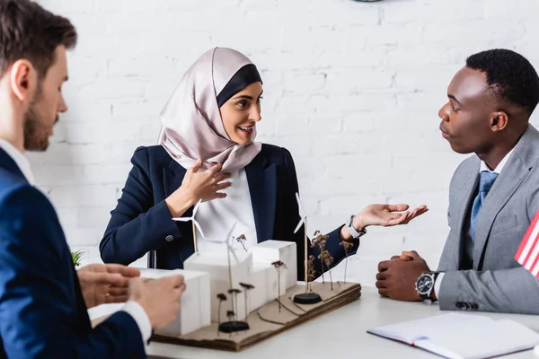 Lächelnde arabische Geschäftsfrau, die mit der Hand zeigt, während sie mit einem afrikanisch-amerikanischen Geschäftspartner in der Nähe eines Ökostrommodells spricht, verschwommener Vordergrund — Stockfoto