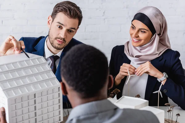 Hombre de negocios señalando con pluma en la construcción de maqueta cerca del modelo de central eléctrica y socios comerciales interracial, borrosa primer plano - foto de stock