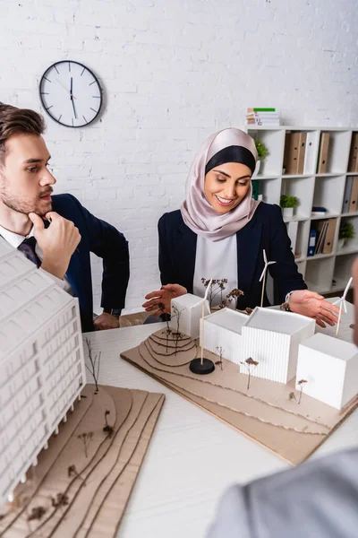 Happy arabian businesswoman pointing at model of green energy station near business partner, blurred foreground — Stock Photo