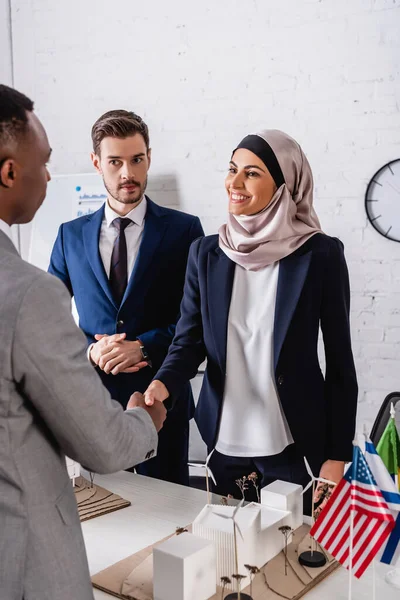 African american businessman shaking hands with happy arabian business partner near maquette of green energy station and international flags — Stock Photo