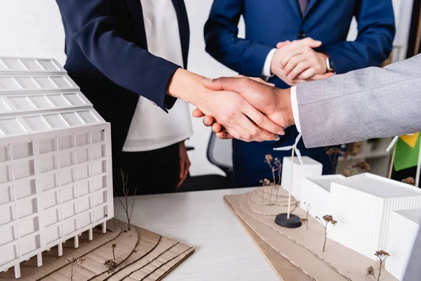 Partial view of interracial business partners shaking hands near model of building with alternative power station — Stock Photo