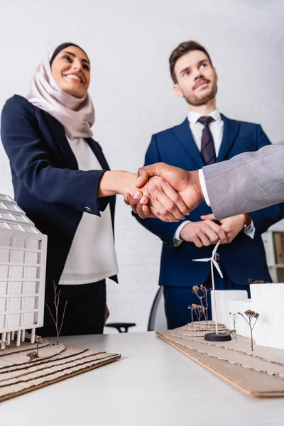 Feliz mujer de negocios árabe estrechando la mano con hombre de negocios afroamericano cerca de modelos de construcción y estación de energía verde, fondo borroso - foto de stock