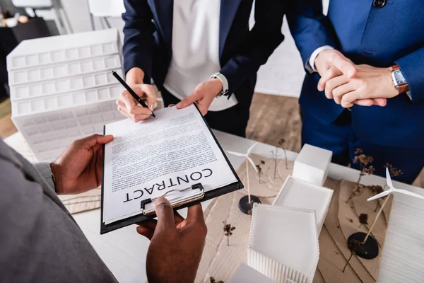 Partial view of woman signing contract near interracial business partners and green power station model — Stock Photo